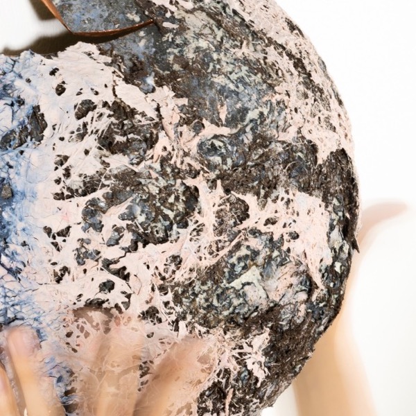 Pleiad. One of Many — the encrusted disc held against the wall in two hands, pink webbing over dark grey